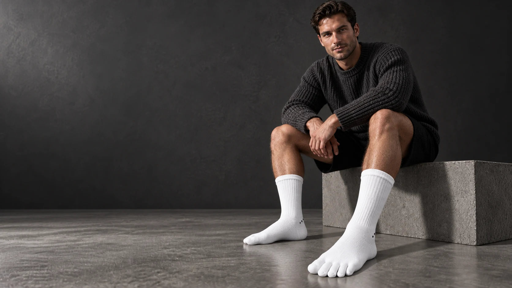 A man in charcoal knitwear and white Albalana toe socks in a dark studio.
